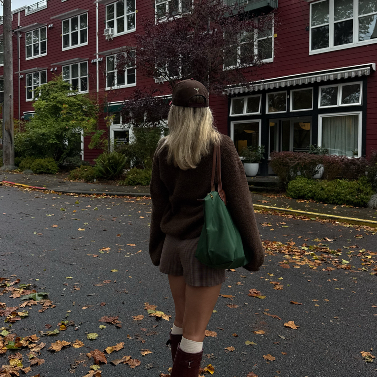 A woman viewed from behind walking on a leaf-strewn street, wearing a brown knit sweater, brown shorts, a brown beanie, white socks, and brown boots, while carrying a large dark green tote bag. A red building with white-framed windows is in the background.