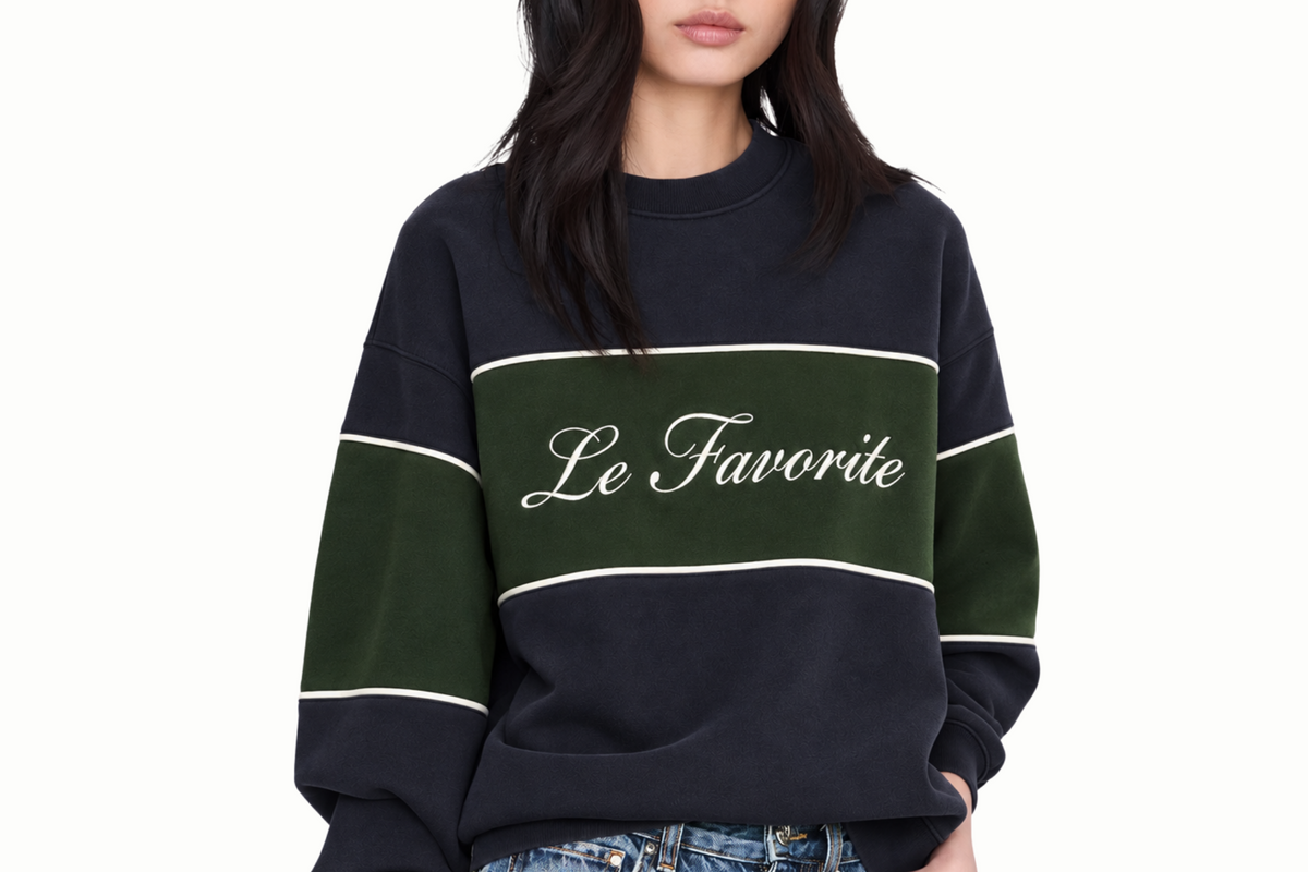 A model wearing a navy blue sweatshirt with a dark green horizontal chest stripe, white piping details, and 'Le Favorite' written in white script across the center. The model is also wearing blue jeans against a plain white background.