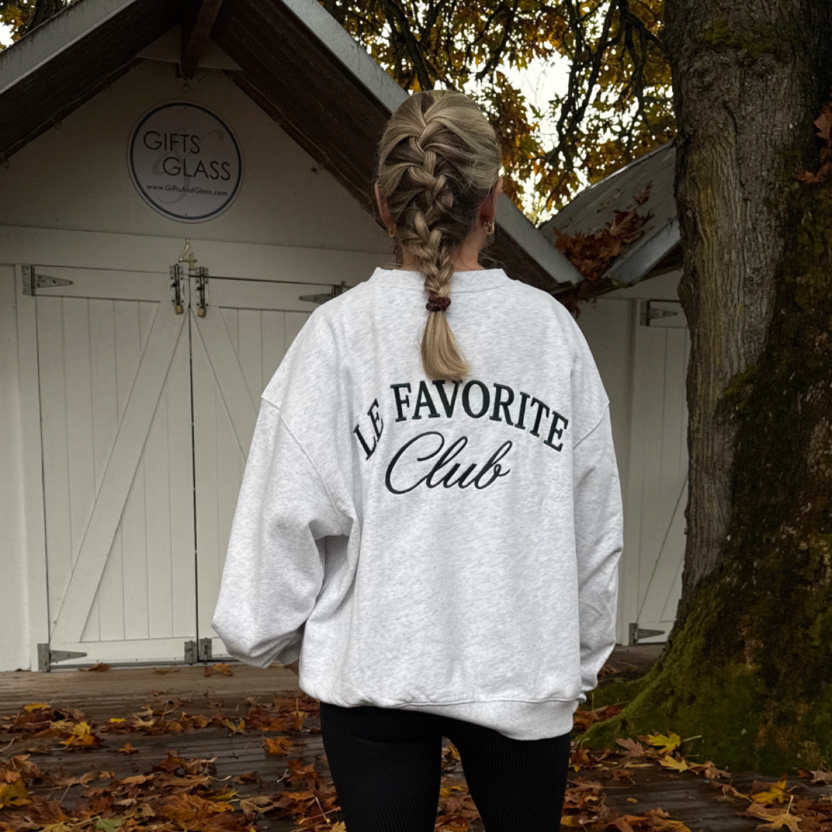 A woman viewed from behind wearing an oversized light gray sweatshirt with 'LE FAVORITE Club' printed in black across the back, standing outdoors in front of a white wooden shed with fallen leaves on the ground.