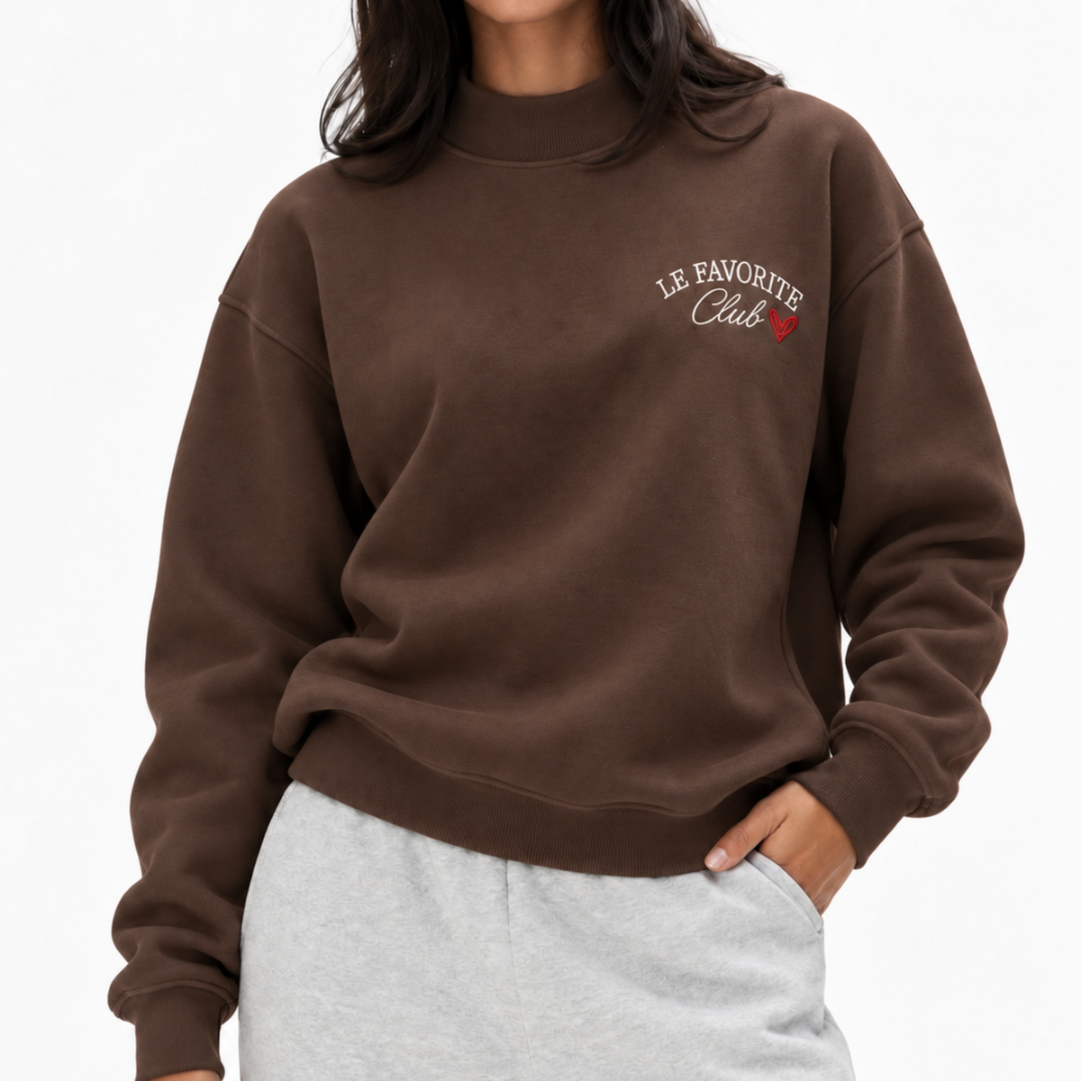 A person wearing a brown crewneck sweatshirt with 'LE FAVORITE Club' embroidered in white and red on the left chest, paired with light gray sweatpants against a plain white background.