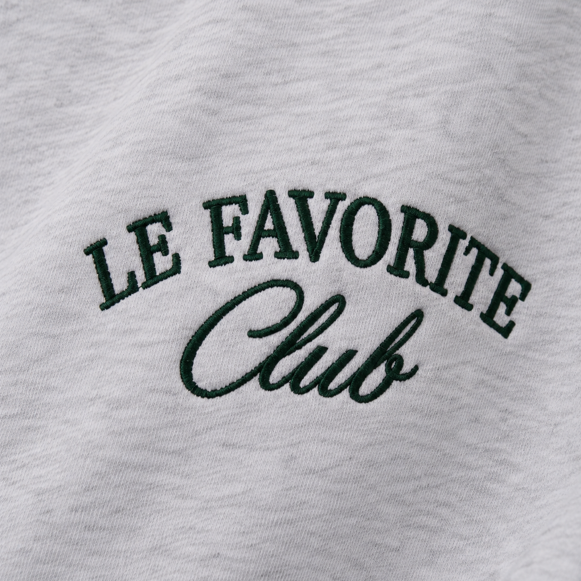 Close-up of a light heather gray sweatshirt featuring dark green embroidered text that reads 'LE FAVORITE Club' across the chest.