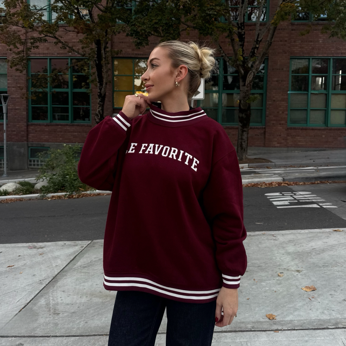 A woman wearing an oversized maroon sweatshirt with white lettering that reads 'E FAVORITE' and white striped ribbed collar, cuffs, and hem, standing outdoors on a sidewalk in front of a brick building.