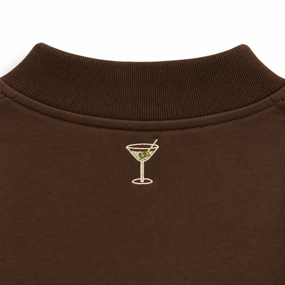Close-up of a dark brown ribbed knit garment, likely a polo shirt collar, featuring a small embroidered cocktail glass with green olives on the chest.