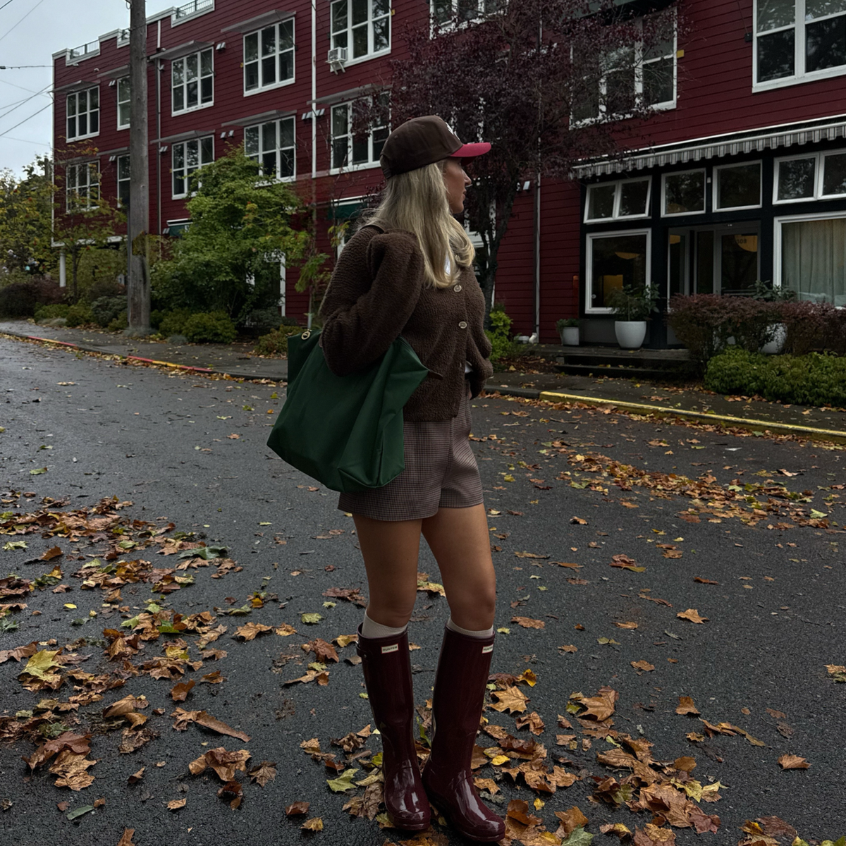 A woman stands on a leaf-strewn street wearing a brown fuzzy sweater, brown shorts, dark brown rain boots, and a brown cap, carrying a large green tote bag. A red building is in the background.